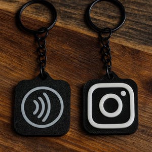 Puede incluir: Dos llaveros negros con una cadena y un anillo negros. Un llavero presenta un símbolo de pago sin contacto blanco, y el otro tiene un logotipo de Instagram blanco. Los llaveros están sobre una superficie de madera.