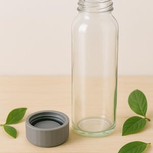 Könnte beinhalten: Eine klare Glasflasche mit grauem Deckel steht auf einer hellen Holzoberfläche neben grünen Blättern. Die Flasche ist zylindrisch und leer. Der Deckel ist ein kurzer Zylinder.