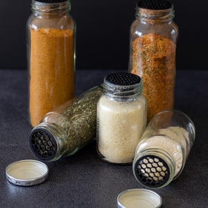 Könnte beinhalten: Vier Gewürzgläser mit schwarzen Streudeckeln, gefüllt mit verschiedenen Gewürzen. Ein Glas enthält ein hellorangefarbenes Pulver, ein anderes eine Kräutermischung und zwei weitere weiße und hellbraune Pulver. Die Gläser stehen auf einer dunklen Oberfläche.