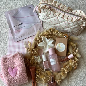 Puede incluir: Una colección de artículos de belleza que incluye una mascarilla de gel de colágeno Medicube, un estuche floral acolchado, un calcetín rosa con corazones, una corona de flores secas, un tónico Glow Recipe, esmalte Essie, aceite Nuxe y un dije de ojo dorado.