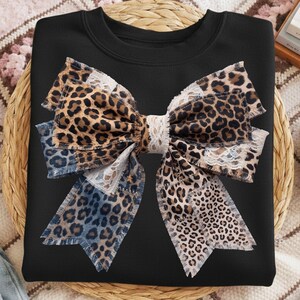 Puede incluir: Sudadera negra con un diseño de lazo grande. El lazo incorpora estampado de leopardo, tela azul tipo denim y detalles de encaje blanco. El lazo es el punto focal, sobre el fondo negro liso de la sudadera.