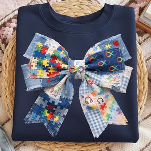 Op de afbeelding: Marineblauw sweatshirt met een groot strikdesign. De strik is gemaakt van denim patches, puzzelstukjes en oneindigheidssymbolen in verschillende kleuren. De strik heeft een gerafelde rand en een kanten rand.