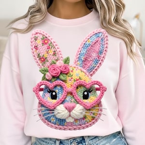 Peut inclure: Un sweat-shirt rose clair orné d'un visage de lapin tricoté coloré avec des lunettes en forme de cœur. Le lapin a des accents floraux sur les oreilles et la tête. Le design incorpore des couleurs rose, bleu, jaune et vert, créant un look joyeux et festif.