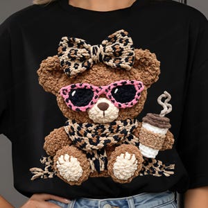 May include: A black t-shirt featuring a brown teddy bear design. The bear wears pink sunglasses, a leopard print bow, and a scarf. It holds a coffee cup with steam rising.