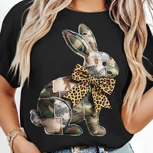Può includere: T-shirt nero con un design a coniglio patchwork. Il coniglio è realizzato in tessuto mimetico e ha un fiocco leopardato. Il design è centrato sulla maglietta, creando un look giocoso e accattivante.