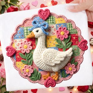 以下が含まれることがあります： オレンジ色のくちばしと足を持つ白いかぎ針編みのガチョウ。青いリボンをつけ、ピンクの花とハートに囲まれています。ガチョウは、ピンクのスカラップボーダーのあるパッチワークの背景に配置されています。
