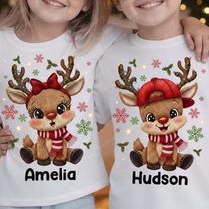 May include: White t-shirts with cartoon reindeer designs. One reindeer wears a red bow, the other a red cap. Both have antlers with lights, red and white striped scarves, and the names "Amelia" and "Hudson" printed below.
