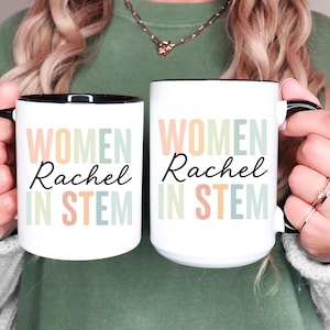 Pode incluir: Duas canecas de cerâmica brancas com interiores e alças pretas. Cada caneca apresenta o texto "WOMEN Rachel IN STEM" em cores pastel. As canecas são seguradas por uma pessoa.