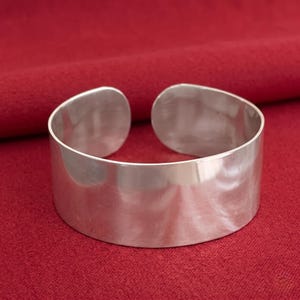 May include: A wide, open-cuff bracelet made of polished silver. The bracelet has a smooth, reflective surface and a simple, modern design. The open ends allow for easy wear. The bracelet is displayed on a red background.