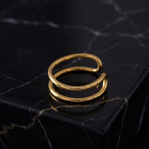 Könnte beinhalten: Ein goldfarbener Ohrklemme mit Doppelband-Design. Das Schmuckstück besteht aus glänzendem, reflektierendem Metall und liegt auf einer schwarzen Marmoroberfläche. Der Ohrklemme umschließt das Ohr und bietet eine moderne, minimalistische Ästhetik.