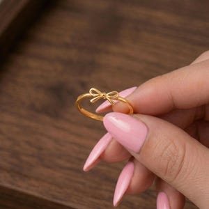 Op de afbeelding: Een goudkleurige ring met een delicaat strikdesign. De ring wordt getoond tegen een achtergrond van een houten oppervlak. De strik is het middelpunt, met zijn ingewikkelde details en gepolijste afwerking. De ring is een sieraad.