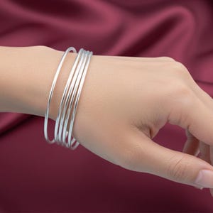Pode incluir: Um conjunto de pulseiras finas e prateadas empilhadas em um pulso. As pulseiras são lisas e brilhantes, refletindo a luz. O fundo é um tecido bordô escuro.