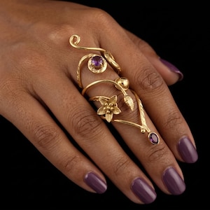 Pode incluir: Um anel dourado para vários dedos com desenhos florais e de folhas, e pequenas pedras preciosas roxas. O anel envolve o dedo com uma estética ornamentada inspirada na natureza. O anel está em uma mão com esmalte de unha roxo.