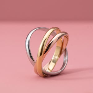 May include: A three-band ring with interlocking bands in silver, gold, and rose gold. The bands are smooth and rounded, creating a modern and elegant design. The ring is set against a soft pink background.
