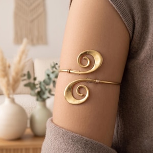 May include: Gold-toned arm cuff featuring a double spiral design. The cuff is worn on the upper arm and has a simple, elegant style. The metal has a matte finish, and the spirals are the focal point of the piece.