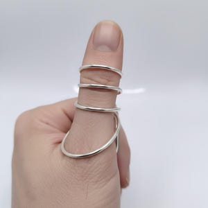 May include: A silver-colored ring designed to be worn on the thumb. The ring features a series of connected, curved bands that wrap around the thumb. The design is modern and minimalist, with a polished finish.