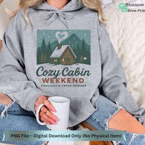 May include: A gray hoodie featuring a graphic of a cozy cabin in a snowy forest with the text "Cozy Cabin Weekend". The design includes a heart-shaped wisp of smoke. The hoodie is a digital download, PNG file only.