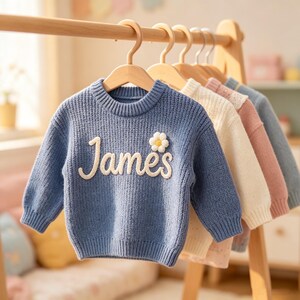 Op de afbeelding: Een blauwe gebreide trui met lange mouwen, voorzien van de naam James in witte cursieve letters en een kleine witte madeliefjesapplicatie op de voorzijde. De trui hangt aan een houten kleerhanger naast andere truien in crème, roze en lichtblauw.