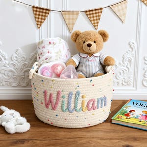 Puede incluir: Una cesta de cuerda color crema con el nombre "William" bordado en hilo colorido. La cesta contiene un osito de peluche, biberones, una manta y un conejito de peluche. Un libro infantil está al lado.