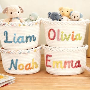 K&ouml;nnte beinhalten: Vier wei&szlig;e Seilk&ouml;rbe mit den gestickten Namen Liam, Olivia, Noah und Emma. Jeder Korb ist mit Pl&uuml;schtieren gef&uuml;llt. Die K&ouml;rbe haben einen gemusterten Rand und stehen auf einer Holzoberfl&auml;che. Eine Lampe und ein Bilderrahmen befinden sich im Hintergrund.