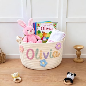 K&ouml;nnte beinhalten: Ein cremefarbener Seilkorb mit dem Namen "Olivia" in bunten Garnbuchstaben und Blumenakzenten. Der Korb enth&auml;lt ein rosa Hasenpl&uuml;schtier, ein Buch und ein wei&szlig;es Handtuch. Zwei Babyrasseln liegen auf dem Boden.