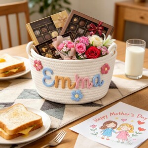 May include: A white rope basket with the name "Emma" in blue, pink, and yellow yarn, decorated with flowers. The basket contains chocolates and a bouquet of pink and red roses. A "Happy Mother's Day" card and a glass of milk are also present.