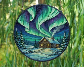 May include: Round stained glass art piece depicting a winter cabin scene under the aurora borealis. The artwork features a cabin with lit windows, snow-covered ground, and green and purple northern lights. The piece is framed in black and hangs from a chain.