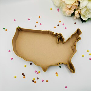 May include: A wooden cookie cutter in the shape of the United States. The cutter is a light brown color and has a detailed outline of the country's borders. Colorful candies and a bouquet of flowers are scattered around the cutter on a white surface.