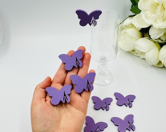 May include: A collection of purple butterfly-shaped wine glass charms. The charms are made of a flat material and have a slit to attach to the glass. A hand holds some of the charms, with others scattered around a wine glass.