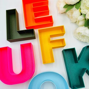 May include: Colorful, 3D-printed letters spelling out "LEFUKQ" in various bright colors. The letters are orange, yellow, pink, blue, and green. The letters are arranged on a white surface, with white flowers in the background.