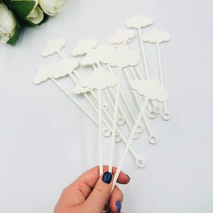 May include: A collection of white cloud-shaped drink stirrers. Each stirrer has a cloud-shaped top and a long, thin stem with a small loop at the end. The stirrers are held in a hand with blue nail polish, with white flowers in the background.