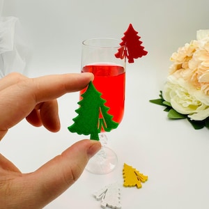 May include: A clear champagne flute filled with red liquid, adorned with a red Christmas tree-shaped drink charm. Additional green, yellow, and white tree-shaped charms are scattered nearby. A hand is holding a green tree charm.