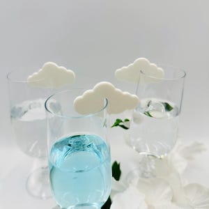 May include: Four clear stemmed glasses, three filled with water and one with blue liquid, each adorned with a white cloud-shaped decoration. White flowers and greenery are at the base of the glasses, creating a whimsical, refreshing aesthetic.