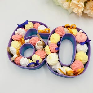 May include: A purple and blue number 60 filled with colorful meringues and tied with ribbons. The meringues are in shades of pink, yellow, and white. The ribbons are blue, gold, and tan. A bouquet of flowers is in the background.