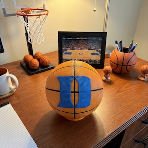 May include: An orange basketball with a large blue "D" logo is the focal point of this image. A miniature basketball hoop, several small basketball figurines, and a basketball-shaped pen holder are also visible on a wooden desk. A framed photo of a basketball court is in the background.