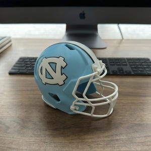 May include: A miniature light blue football helmet with a white facemask and the University of North Carolina logo. The helmet is on a wooden desk with a computer and keyboard in the background.