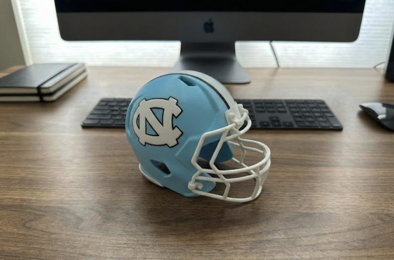 May include: A miniature light blue football helmet with a white facemask and the University of North Carolina logo. The helmet is on a wooden desk with a computer and keyboard in the background.