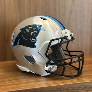 May include: Silver football helmet with a black facemask and a blue stripe. The helmet features a black and blue panther logo on the side. The helmet is sitting on a wooden surface.