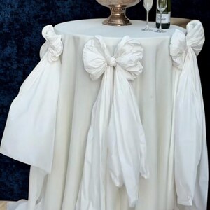 May include: A white round table with a draped tablecloth and large bow accents. A silver champagne bucket holds bottles of champagne, with two glasses and a bottle on the table. The table is set against a dark blue backdrop.