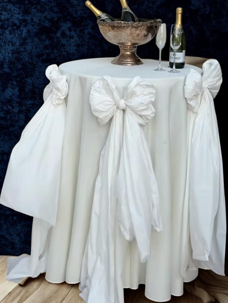 May include: A white round table with a draped tablecloth and large bow accents. A silver champagne bucket holds bottles of champagne, with two glasses and a bottle on the table. The table is set against a dark blue backdrop.