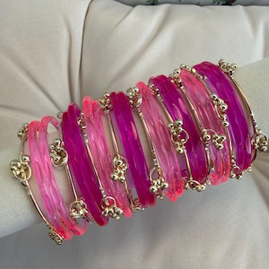 Könnte beinhalten: Ein Stapel durchscheinender Armreifen in verschiedenen Rosa- und Magentatönen. Jeder Armreif hat eine texturierte, facettierte Oberfläche und ist mit einem kleinen, goldfarbenen Metallanhänger aus drei winzigen Kugeln verziert.
