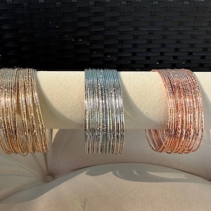 May include: Three sets of thin, textured bangle bracelets displayed on a horizontal stand. The set on the left is gold, the centre set is silver, and the set on the right is rose gold. Each bangle features a fine, glittery, metallic finish.