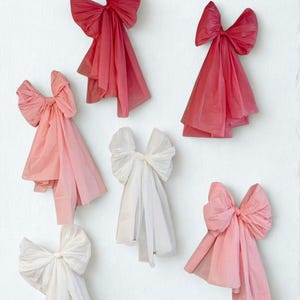 May include: An assortment of decorative bows in various shades of pink and white are arranged against a white background. The bows are made of a soft, flowing material, with a layered design. The bows are in different sizes and shapes.