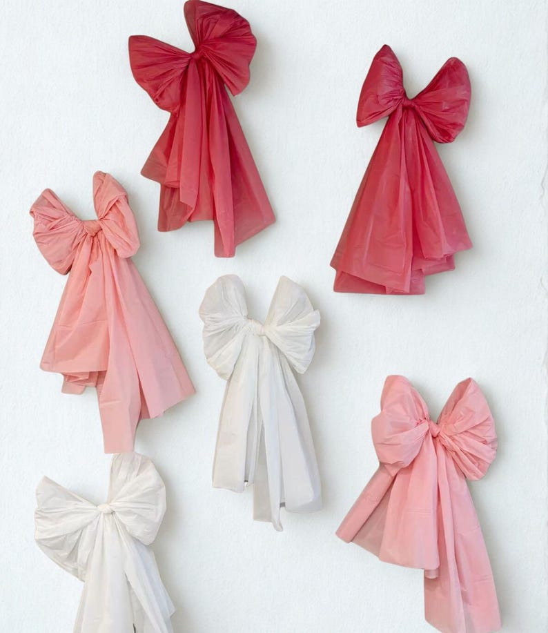 May include: An assortment of decorative bows in various shades of pink and white are arranged against a white background. The bows are made of a soft, flowing material, with a layered design. The bows are in different sizes and shapes.