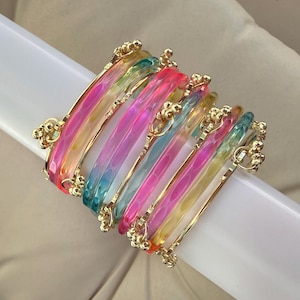 Pode incluir: Um conjunto de pulseiras translúcidas e multicoloridas com um gradiente de tons rosa, azul, amarelo e verde. Cada pulseira é acentuada com finas faixas de metal dourado e pequenos grupos decorativos de metal dourado nos pontos de união.