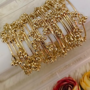 May include: A collection of gold-colored bangles. Each bangle features a series of small, round, decorative elements. The bangles are arranged in a clear container, with a few flowers in the background.