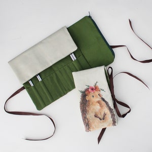 Könnte beinhalten: Eine grüne und beige Stoff-Bleistiftrolle mit mehreren Fächern zum Aufbewahren von Bleistiften oder anderen Kunstmaterialien. Die Rolle wird mit einem Reißverschluss geschlossen und hat ein braunes Band. Eine separate beige Stofftasche mit einem Igel-Design ist ebenfalls enthalten.