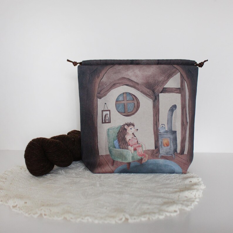 K&ouml;nnte beinhalten: Eine braune Kordeltasche mit einer Aquarell-Illustration eines Igels, der in einem Stuhl neben einem Kamin sitzt. Die Illustration spielt in einem gem&uuml;tlichen H&auml;uschen mit einem Fenster und einem Bild an der Wand.