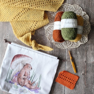May include: A still-life image featuring crochet and knitting supplies. A yellow crocheted scarf, three skeins of yarn in orange, green, and yellow, a crochet hook, and a white drawstring bag with an otter illustration are arranged on a wooden surface.