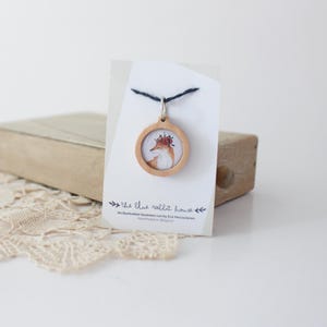Pode incluir: Um colar com um pingente de madeira redondo com uma ilustração de uma raposa. O pingente está suspenso em uma corrente prateada. O cartão diz "the blue rabbit house" e "An illustration business run by Eva Vercauteren. Handmade in Belgium."